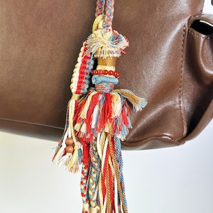 Pode incluir: Um pendente decorativo para mala com um arranjo complexo de fios trançados e soltos em tons de vermelho, azul, creme e bege. O acessório inclui pequenas contas esféricas de madeira e vermelhas integradas no design, pendurado numa mala de pele castanha.