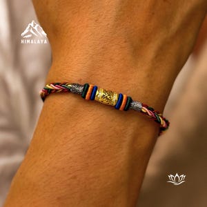 Peut inclure: Bracelet tressé multicolore avec une perle centrale dorée ornée d'un motif complexe. Le bracelet est composé de fils rouges, jaunes, verts, bleus et noirs. Le mot "HIMALAYA" est visible en haut à gauche.