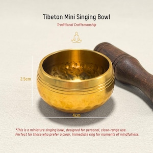 May include: A small, golden Tibetan singing bowl, 4 cm in diameter and 2.5 cm high, with a wooden mallet. The bowl has a textured surface and is labeled "Tibetan Mini Singing Bowl" and "Traditional Craftsmanship."