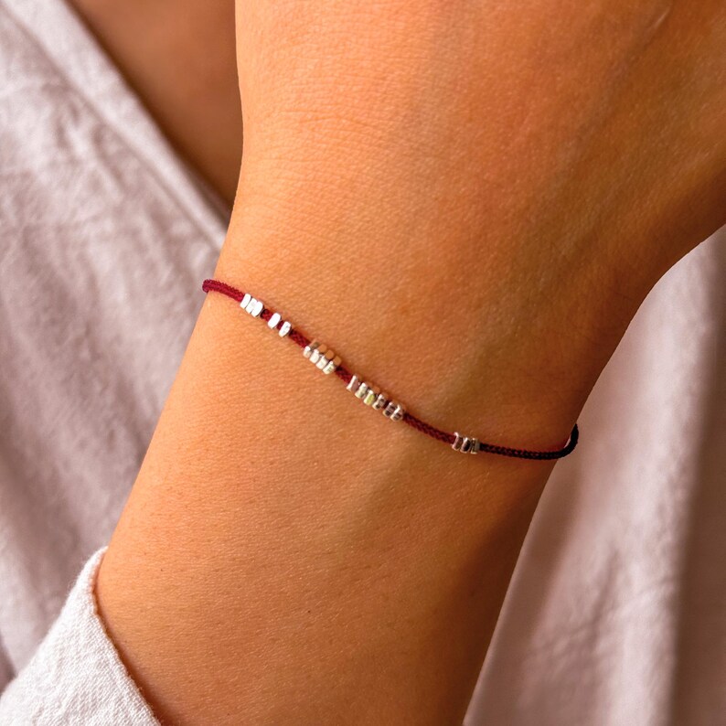 May include: A maroon string bracelet with small silver and tan beads. The bracelet is worn on a wrist, and the beads are evenly spaced along the string. The bracelet is a delicate piece of jewelry.
