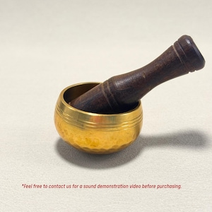 May include: A small, golden singing bowl with a dark brown wooden striker. The bowl has a hammered texture and a polished finish. The striker is angled across the bowl. The text at the bottom says "Feel free to contact us for a sound demonstration video before purchasing."