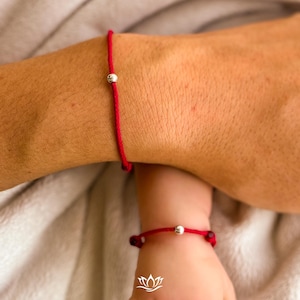May include: Two red string bracelets, one on an adult wrist and the other on a child's wrist. Each bracelet features a small, silver-colored bead. The bracelets are set against a soft, neutral-toned background.