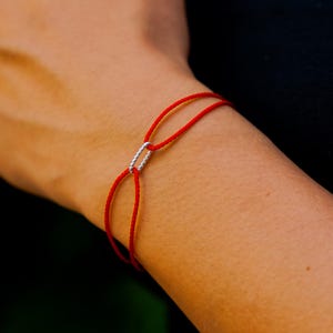 May include: A red string bracelet with a silver-colored rectangular clasp. The bracelet is worn on a wrist, with the focus on the jewelry. The background is dark, emphasizing the bracelet's design.