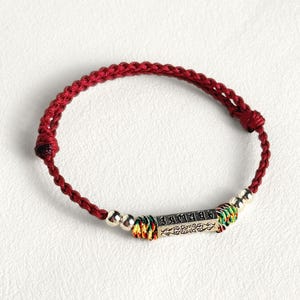 May include: A red braided bracelet with a silver rectangular charm. The charm has etched text and is flanked by silver beads and colorful woven accents. The bracelet is on a white background.