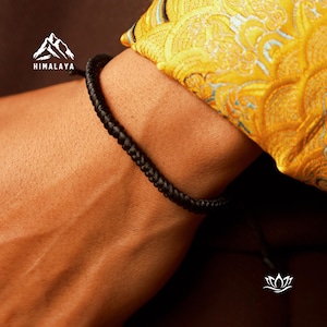 May include: A black braided bracelet on a person's wrist. The bracelet is made of a thick, durable cord and has a simple, elegant design.