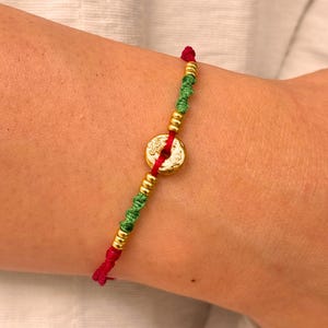 May include: A bracelet featuring a central gold-coloured circular charm with an embossed pattern, flanked by small gold-coloured beads and sections of green knotted cord, all strung on a vibrant red cord with a visible knot at the top.
