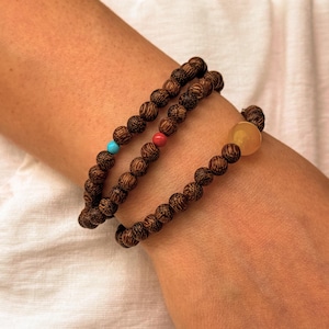May include: Three wooden beaded bracelets are wrapped around a wrist. The bracelets are made of dark brown, round wooden beads. One bracelet has a turquoise bead, another has a red bead, and the third has a yellow bead.