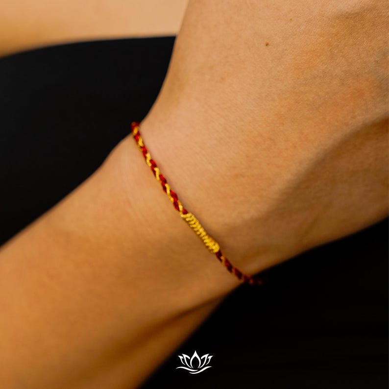 Puede incluir: Primer plano de una pulsera trenzada roja y dorada. La pulsera se lleva en la mu&ntilde;eca y presenta una banda central dorada. El fondo es de color oscuro y s&oacute;lido.