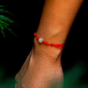 May include: A red string bracelet with a light-colored, round bead. The bracelet is tied with knots and worn on the wrist. The background is dark, with a hint of green foliage.