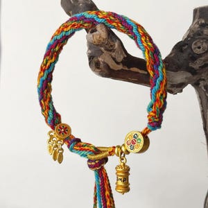 Pu&ograve; includere: Un braccialetto intrecciato colorato con dettagli dorati. Il braccialetto presenta un arcobaleno di colori, tra cui rosso, giallo, blu e verde. Ciondoli dorati e una chiusura decorativa aggiungono dettagli.