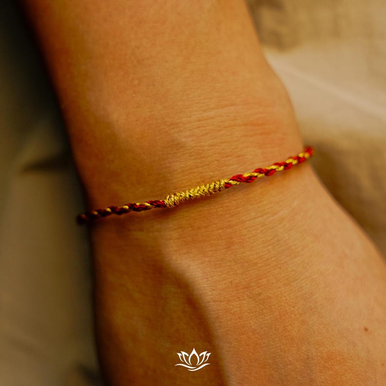 Puede incluir: Primer plano de una pulsera trenzada roja y dorada. La pulsera tiene una secci&oacute;n central dorada. La pulsera se lleva en la mu&ntilde;eca. El fondo es de color beige neutro.