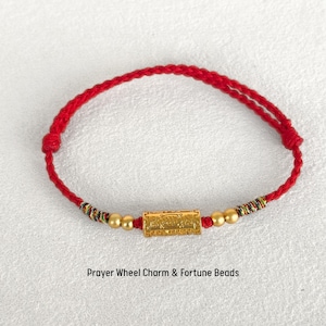 May include: A red braided cord bracelet featuring a central gold-colored cylindrical prayer wheel charm, flanked by two small gold-colored beads on each side and multi-colored thread accents. The text Blessing Bracelet and Prayer Wheel Charm & Fortune Beads is visible.