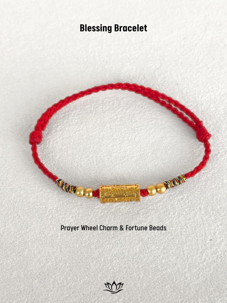 May include: A red braided cord bracelet featuring a central gold-colored cylindrical prayer wheel charm, flanked by two small gold-colored beads on each side and multi-colored thread accents. The text Blessing Bracelet and Prayer Wheel Charm & Fortune Beads is visible.