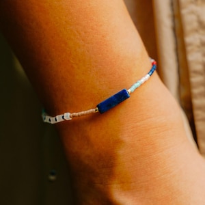 Op de afbeelding: Een kralenarmband met een levendige mix van kleuren. De armband heeft een donkerblauwe rechthoekige kraal als middelpunt, aangevuld met kleinere kralen in wit, beige, lichtblauw en rood. De armband is een delicaat sieraad.