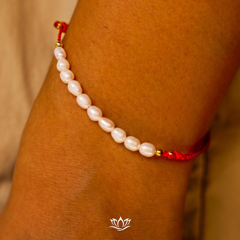 Peut inclure: Un bracelet d&eacute;licat compos&eacute; de perles blanches. Les perles sont enfil&eacute;es sur un cordon rouge, avec des d&eacute;tails dor&eacute;s. Le bracelet est pr&eacute;sent&eacute; sur un poignet, mettant en valeur son design &eacute;l&eacute;gant.