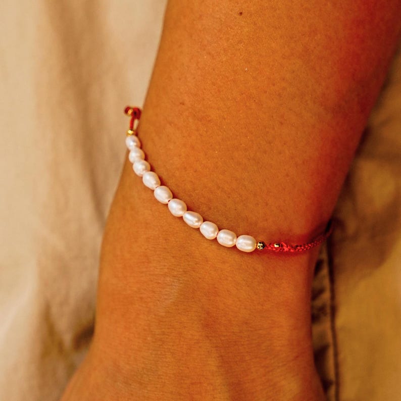 Peut inclure: Bracelet d&eacute;licat orn&eacute; de perles blanches. Les perles sont enfil&eacute;es sur un cordon rouge, avec des d&eacute;tails dor&eacute;s. Le bracelet est port&eacute; sur un poignet, sur un fond neutre.