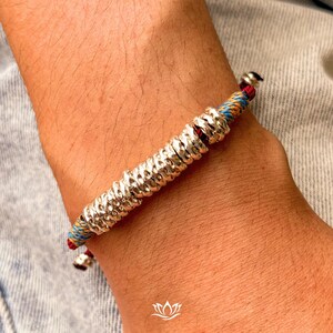 May include: Silver bracelet with a textured, cylindrical centre and colourful woven cord accents. The bracelet features a series of small, circular silver elements. The cord includes red, blue, and orange threads. The bracelet is worn on a wrist.