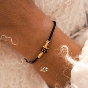 May include: A black string bracelet with a gold charm featuring a small seated figure. The charm is adorned with intricate details and a small bead.
