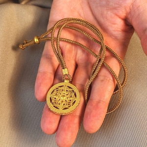 Pode incluir: Colar com pingente dourado com um design circular de filigrana. O pingente pende de um cordão trançado. O colar é segurado em uma mão contra um fundo neutro. O pingente tem um intrincado padrão vazado.