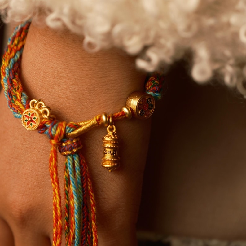 Pu&ograve; includere: Un braccialetto intrecciato colorato con dettagli dorati. Il braccialetto presenta un ciondolo dorato con una piccola campana ed elementi decorativi dorati. I fili intrecciati sono nelle tonalit&agrave; di arancione, giallo, blu e rosso.