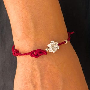 Könnte beinhalten: Ein dunkelrotes Kordelarmband mit einem silberfarbenen Blumenanhänger in der Mitte, zwei kleinen silberfarbenen Kugeln und einem dekorativen Knoten. Das Armband wird mit einem einfachen Knoten an der dünnen, geflochtenen roten Kordel verschlossen.