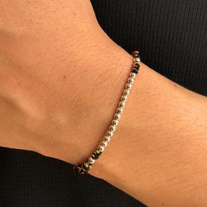 Peut inclure: Un bracelet de perles argentées avec un détail tissé noir et doré. Le bracelet est composé de petites perles rondes argentées. Le détail tissé incorpore des fils noirs, dorés et rouges. Le bracelet est porté sur un poignet.