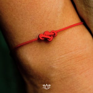 Pode incluir: Um close-up de uma pulseira de fio vermelho com um design de n&oacute; duplo. A pulseira &eacute; feita de um cord&atilde;o fino e tecido. O n&oacute; &eacute; o ponto focal, e a pulseira &eacute; usada no pulso.
