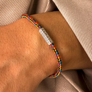 May include: A multi-coloured braided bracelet with red, yellow, blue, and black threads. The bracelet has a silver-coloured rectangular clasp with engraved text. The bracelet is worn on a wrist, with a neutral-coloured shirt in the background.