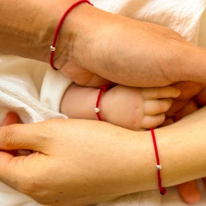 Può includere: Primo piano di tre braccialetti di filo rosso con una piccola perla d'argento. I braccialetti sono indossati sui polsi di tre persone, tra cui un bambino. L'immagine trasmette un senso di famiglia e connessione.