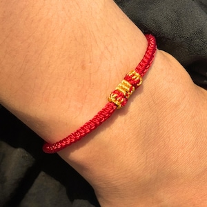 Peut inclure: Un bracelet rouge tressé avec des accents dorés. Le bracelet est fait d'un cordon rouge étroitement tissé et comporte des nœuds décoratifs dorés. Le bracelet est porté sur un poignet.
