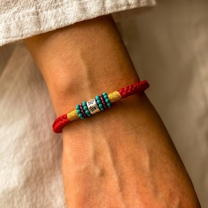 Può includere: Braccialetto rosso intrecciato con dettagli dorati e perline turchesi e rosse. Un cilindro d'argento con caratteri incisi è centrato sul braccialetto. Il braccialetto è indossato al polso.
