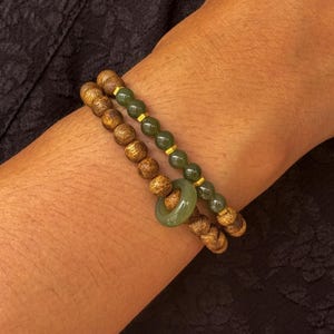 May include: Two bracelets stacked on a wrist. One bracelet features wooden beads, while the other has green beads with gold accents. A green jade ring is on the wooden bead bracelet. The bracelets are a natural, earthy accessory.