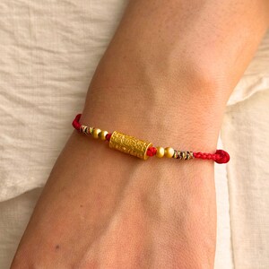 May include: A red cord bracelet featuring a central cylindrical gold bead with intricate embossed patterns, flanked by two smaller spherical gold beads on each side, and finished with decorative multi-colored thread accents near the knots.