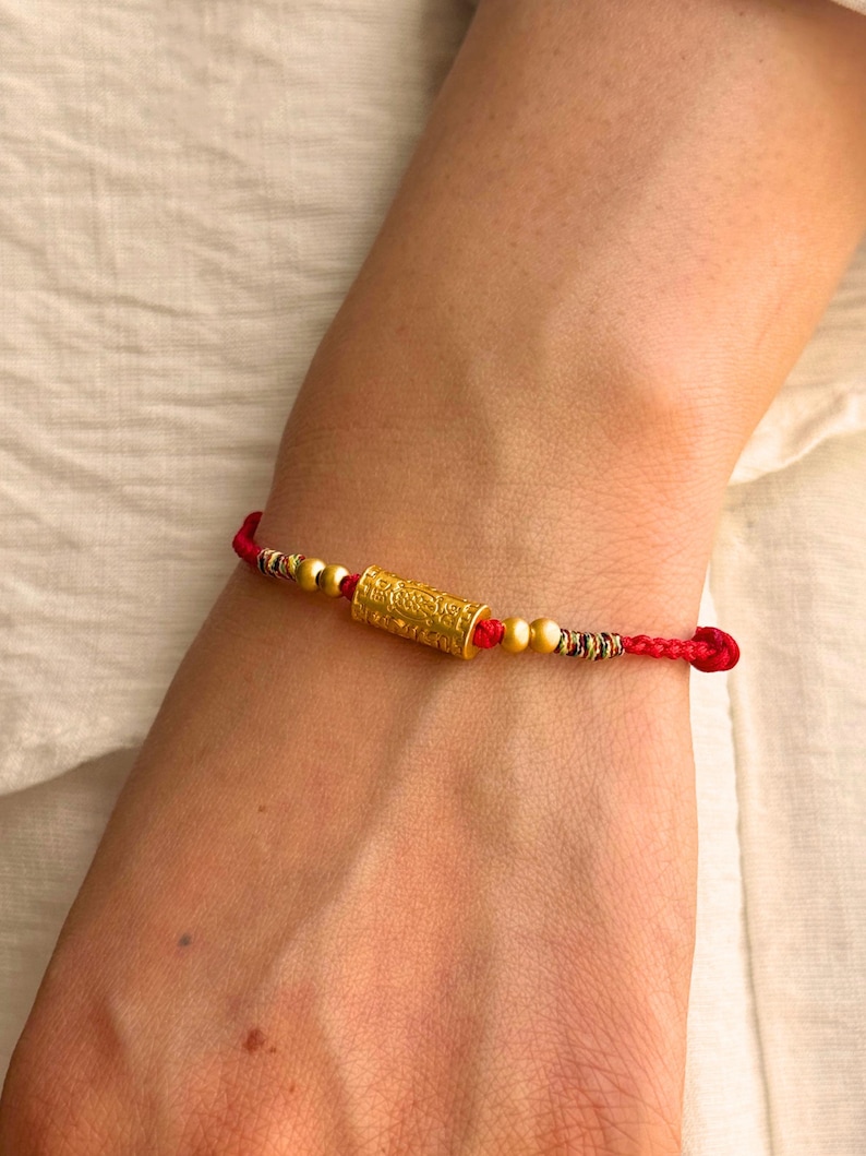 May include: A red cord bracelet featuring a central cylindrical gold bead with intricate embossed patterns, flanked by two smaller spherical gold beads on each side, and finished with decorative multi-colored thread accents near the knots.
