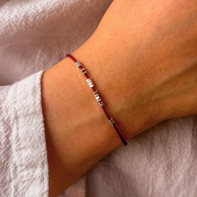 May include: A minimalist bracelet featuring a deep red cord with small, silver-colored rectangular beads. The bracelet is designed to be worn on the wrist, showcasing a simple yet elegant aesthetic. The cord is thin and the beads are evenly spaced.