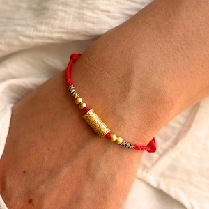 Puede incluir: Una pulsera de cordón rojo con una cuenta central cilíndrica de tono dorado con patrones en relieve, flanqueada por dos cuentas esféricas doradas más pequeñas y dos cuentas de acento oscuras y texturizadas a cada lado.