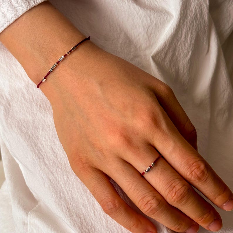 May include: A red string bracelet and ring set. The bracelet features small silver beads, and the ring has a similar design. The jewelry is displayed on a person's hand, against a white background.