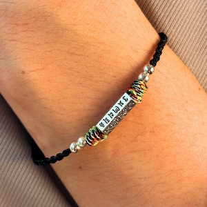May include: Black braided bracelet with a silver rectangular charm. The charm has white lettering and is accented with colorful thread and silver beads. The bracelet is worn on a wrist.