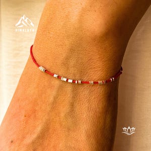 May include: A red string bracelet featuring small silver beads and square details. The bracelet is displayed on a wrist, highlighting its delicate design and craftsmanship. This bracelet is a simple yet elegant piece of jewellery.