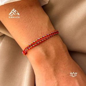Può includere: Un braccialetto intrecciato rosso e multicolore. Il braccialetto è avvolto attorno a un polso. Il braccialetto ha fili rossi, gialli, verdi, blu e bianchi. Il braccialetto è annodato all'estremità. L'immagine ha la parola "HIMALAYA" e un logo di montagna.