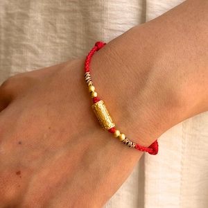 May include: A red braided cord bracelet featuring a central cylindrical gold-toned bead with intricate embossed patterns, flanked by two smaller spherical gold-toned beads and accented with small sections of multi-colored thread detailing.