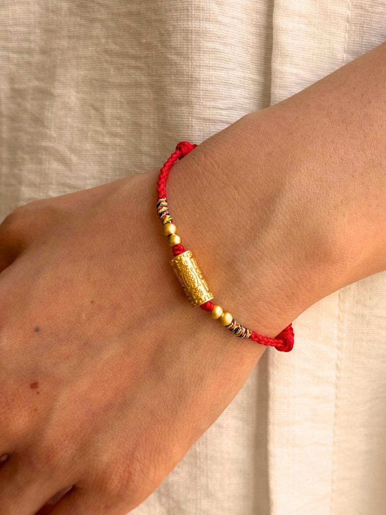 May include: A red braided cord bracelet featuring a central cylindrical gold-toned bead with intricate embossed patterns, flanked by two smaller spherical gold-toned beads and accented with small sections of multi-colored thread detailing.