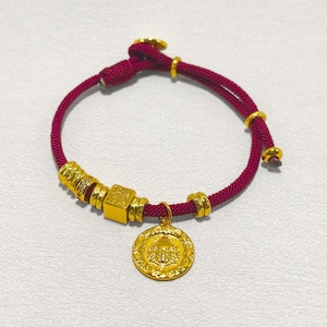 May include: A burgundy cord bracelet with gold-colored accents and a circular pendant. The bracelet features a series of gold beads and a square charm. The pendant has a detailed design. The bracelet is on a white surface.