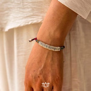 May include: A silver bracelet with a woven design. The bracelet has a central band of tightly wound silver-coloured metal, with colourful threads in blue, green, and red. It is secured with a red and black cord.