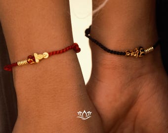 May include: Two bracelets, one red and one black, are worn on wrists. The red bracelet has gold-colored beads and a woven design. The black bracelet also has gold-colored beads and a woven design. The bracelets are likely handmade.
