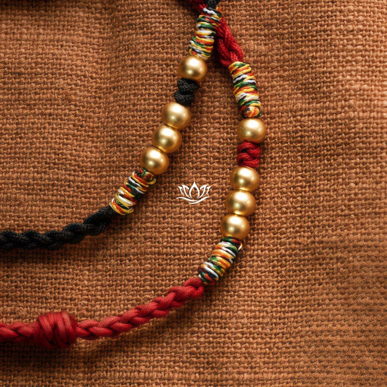May include: A close-up of a red, black, and white braided cord necklace with gold beads. The necklace is lying on a brown fabric surface.