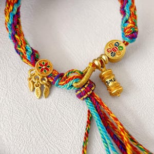 Pu&ograve; includere: Primo piano di un braccialetto intrecciato colorato con abbellimenti color oro. Il braccialetto presenta fili rossi, gialli, blu e verdi. I ciondoli color oro includono un motivo floreale e un piccolo cilindro inciso.