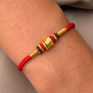 Pode incluir: Uma pulseira de cordão vermelho com uma conta central dourada em forma de barril com símbolos em relevo. O design inclui detalhes decorativos de fio metálico dourado, cordão preto e segmentos de cordão vermelho ao lado da conta central.