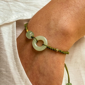 May include: A jade bracelet with a circular pendant and a round bead, strung on a green cord with gold accents. The bracelet is adjustable and has a sliding knot closure. The jade has a pale green colour.