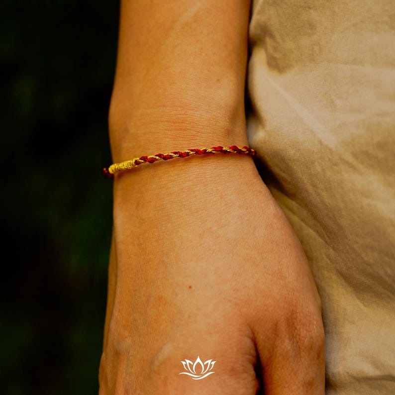 Puede incluir: Una pulsera trenzada con hilos rojos y dorados se lleva en la mu&ntilde;eca. La pulsera est&aacute; hecha de hebras muy tejidas, creando una apariencia texturizada. El fondo est&aacute; borroso, enfocando la atenci&oacute;n en la joya.